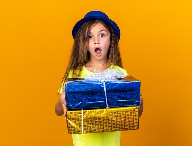 piccola ragazza caucasica emozionante con il cappello blu del partito che tiene i contenitori di regalo isolati sulla parete arancio con lo spazio della copia