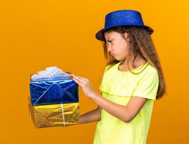 piccola ragazza caucasica scontenta con cappello da festa blu che tiene in mano e guarda scatole regalo isolate sulla parete arancione con spazio copia