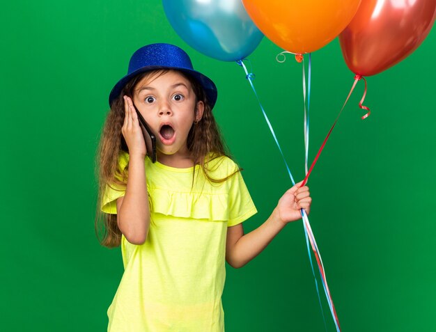 piccola ragazza caucasica sorpresa con cappello blu del partito che tiene palloncini di elio e parla al telefono isolato sulla parete verde con lo spazio della copia