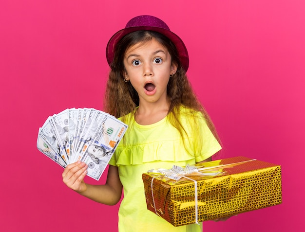 piccola ragazza caucasica sorpresa con cappello da festa viola che tiene scatola regalo e denaro isolato sulla parete rosa con spazio per la copia