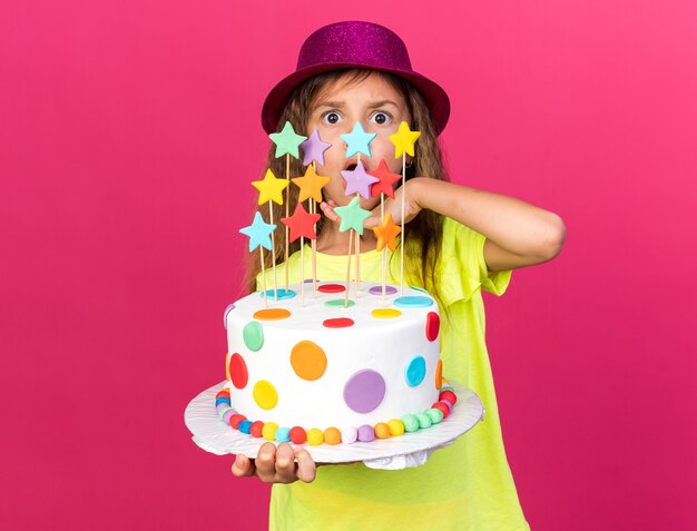 piccola ragazza caucasica sorpresa con il cappello viola del partito che tiene la torta di compleanno e che mette la mano sul mento isolato sulla parete rosa con lo spazio della copia
