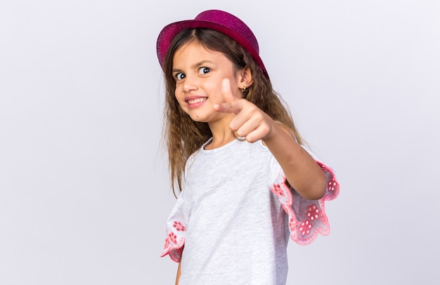 piccola ragazza caucasica sorridente con il cappello viola del partito che indica isolato sulla parete bianca con lo spazio della copia