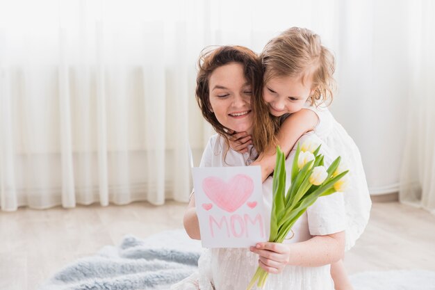 Piccola ragazza che abbraccia la sua mamma dalla parte posteriore mentre sorride la cartolina d&#39;auguri e fiori della tenuta della madre a casa