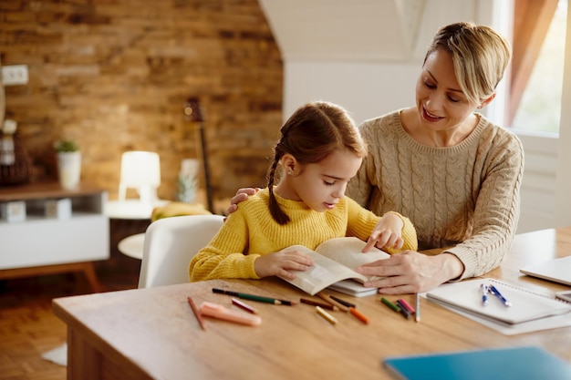 Piccola ragazza che legge un libro mentre impara con l'aiuto di sua madre a casa