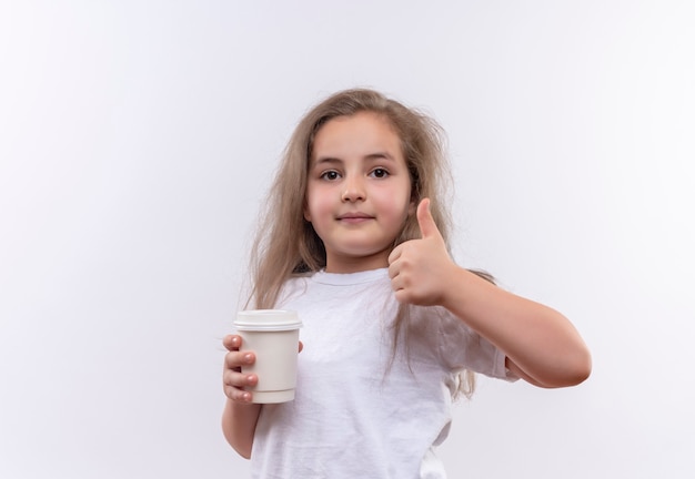 piccola ragazza della scuola che indossa la maglietta bianca che tiene la tazza di caffè il suo pollice in su sulla parete bianca isolata