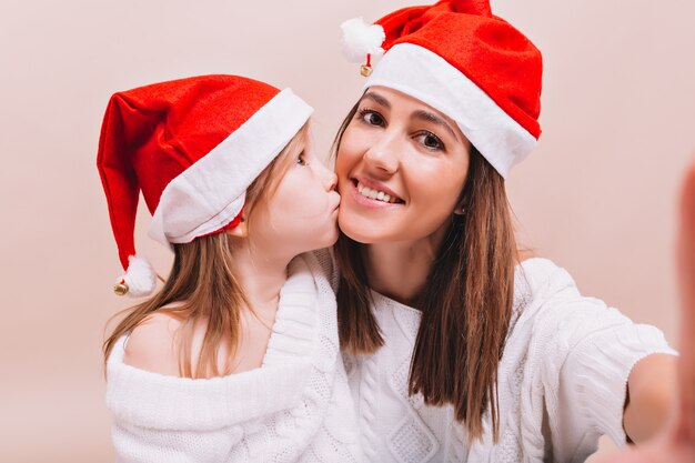 Piccola ragazza graziosa che bacia la sua sorella maggiore che indossa i cappelli di Babbo Natale simili, la donna che fa selfie con sua figlia sul muro isolato, veramente emozioni