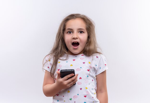 Piccola ragazza sorpresa della scuola che indossa la bocca aperta del telefono della tenuta della maglietta bianca su fondo bianco isolato