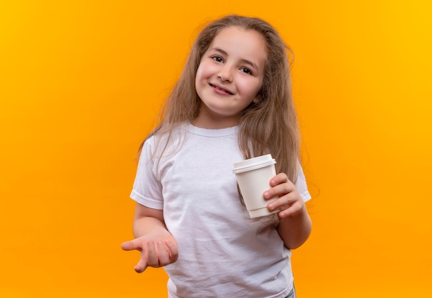 Piccola ragazza sorridente della scuola che indossa la maglietta bianca che tiene tazza di caffè su fondo arancio isolato