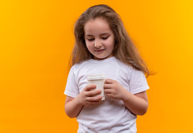 Piccola ragazza sorridente della scuola che indossa la maglietta bianca che tiene tazza di caffè su fondo arancio isolato