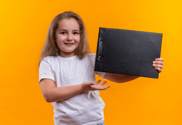 Piccola ragazza sorridente della scuola che porta la lavagna per appunti bianca della tenuta della maglietta su fondo arancio isolato