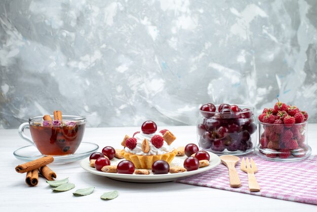 piccola torta cremosa con lamponi, ciliegie e biscotti piccoli, tè alla cannella sulla scrivania a luce bianca, torta alla frutta, crema ai frutti di bosco