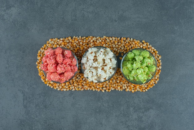 Piccole ciotole da portata di popcorn assortiti circondate da chicchi di mais su fondo marmo. Foto di alta qualità