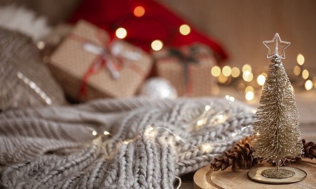 Piccolo albero di Natale lucido decorativo in primo piano su uno sfondo sfocato di una sciarpa lavorata a maglia, decorazioni natalizie e luci bokeh copia spazio.