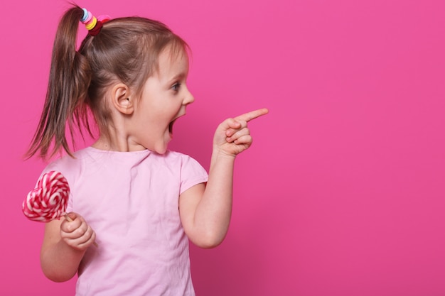 piccolo bambino che apre la bocca ampiamente, guardando dall'altra parte con eccitazione, tenendo il cuore lecca-lecca brillante. La bambina allegra e bionda allegra trascorre felicemente il tempo libero.