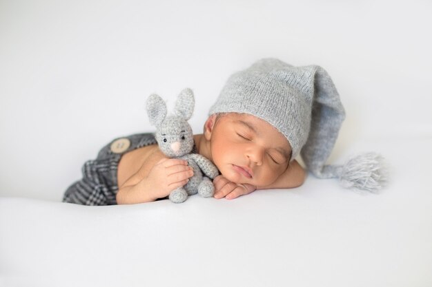 Piccolo bambino che dorme con cappello grigio carino e con coniglio giocattolo nelle sue mani