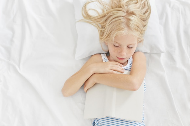 Piccolo bambino con un bell'aspetto che dorme dopo aver letto storie interessanti nel letto, tenendo il libro in mano, sdraiato sul cuscino bianco e vestiti da letto, avendo sogni piacevoli. Leggere prima di dormire