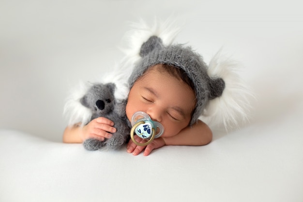 Piccolo bambino di riposo sveglio del neonato appena nato con il cappello grigio e l'orso grigio del giocattolo in sue mano e tettarella sulla sua bocca su un pavimento bianco