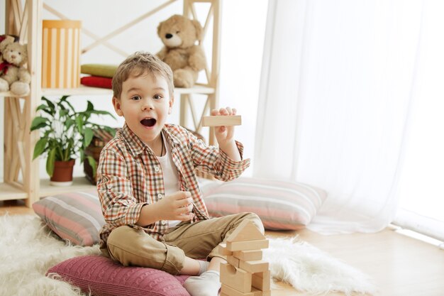 Piccolo bambino seduto sul pavimento. Ragazzo sorpreso abbastanza sorridente che gioca con i cubi di legno a casa. Immagine concettuale con copia o spazio negativo e mock-up per il tuo testo.