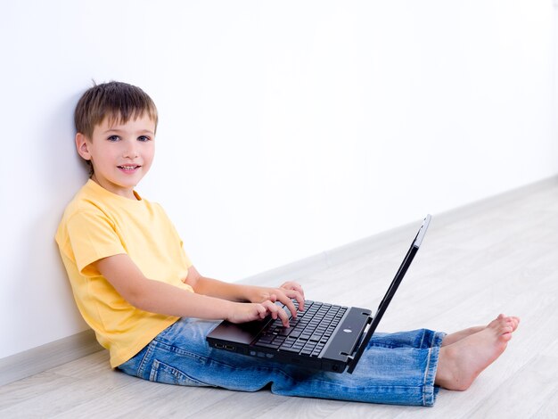 Piccolo bambino sorridente con il computer portatile che si siede nella stanza vuota - all'interno