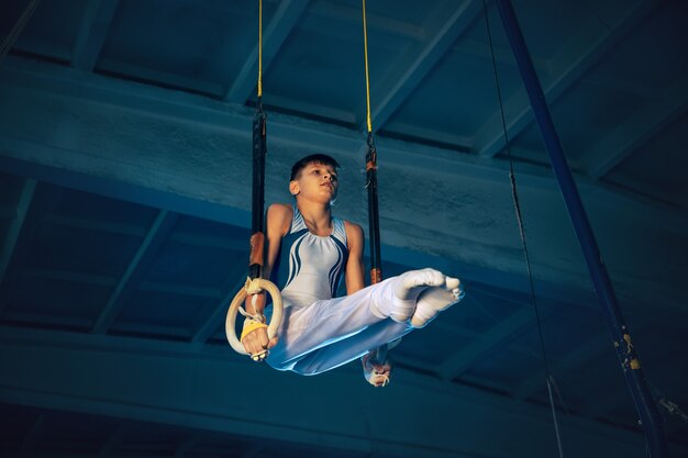 Piccolo ginnasta maschio allenamento in palestra, flessibile e attivo. Ragazzo in forma caucasica, atleta in abiti sportivi bianchi che praticano esercizi per l'equilibrio sugli anelli. Movimento, azione, movimento, concetto dinamico.