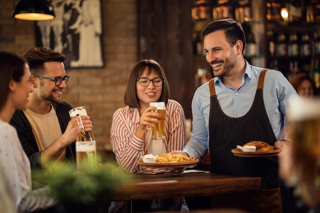 Piccolo gruppo di amici felici che bevono birra mentre il cameriere serve loro uno spuntino in una taverna