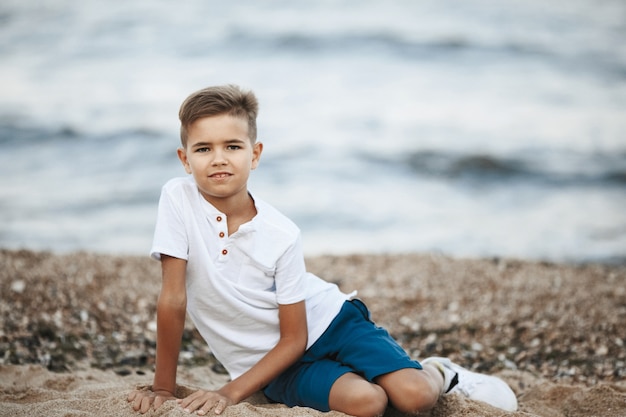 Piccolo ragazzo caucasico è seduto sulla spiaggia vicino al mare e guardando dritto vestito in abiti casual