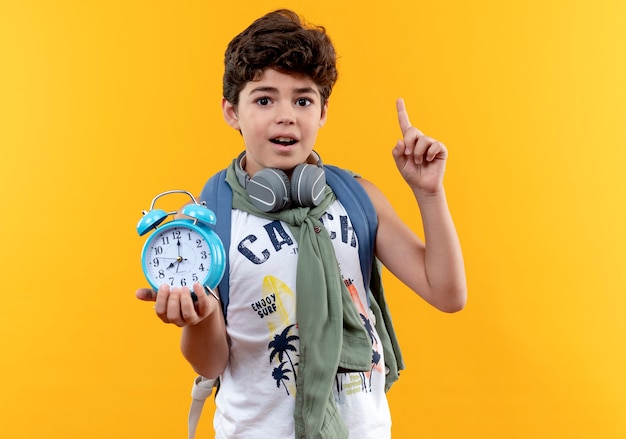 Piccolo scolaro impressionato che indossa borsa posteriore e cuffie che tengono sveglia e indica in alto isolato su sfondo giallo