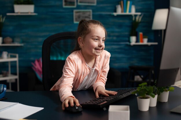 Piccolo scolaro intelligente seduto al tavolo della scrivania usando il computer