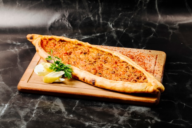 Pide turco tradizionale con carne ripiena, limone e prezzemolo su una tavola quadrata in legno.