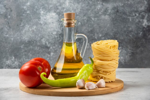 Pila di nidi di pasta cruda, bottiglia di olio d'oliva e verdure sul piatto di legno.