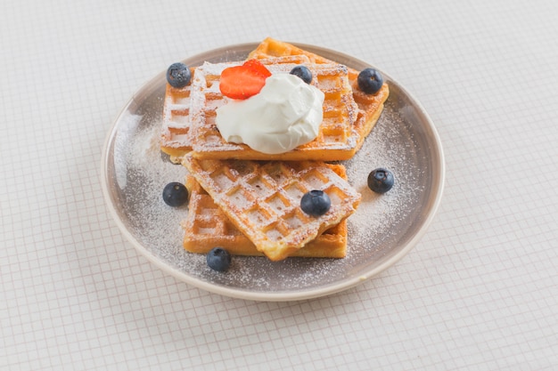 Pila di waffle guarniti con fragole; mirtillo e panna montata sul piatto sopra la tovaglia