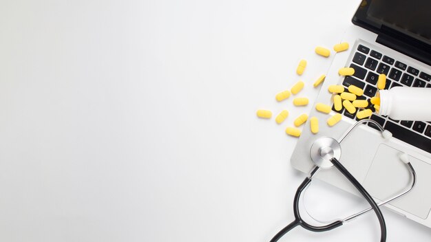 Pillole e stetoscopio gialli rovesciati sul computer portatile sopra fondo bianco