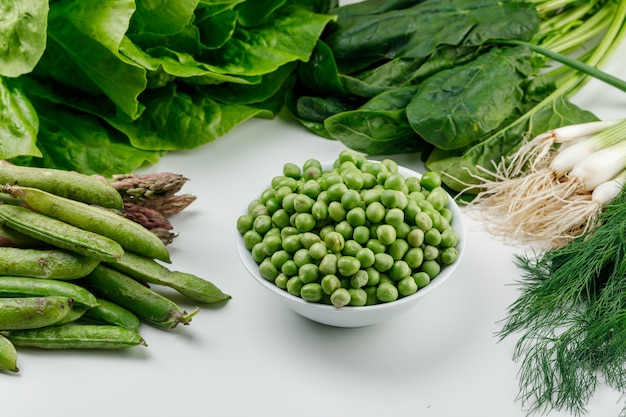 Piselli in una ciotola con baccelli verdi, acetosa, aneto, lattuga, asparagi, cipolle verdi vista dall'alto su un muro bianco