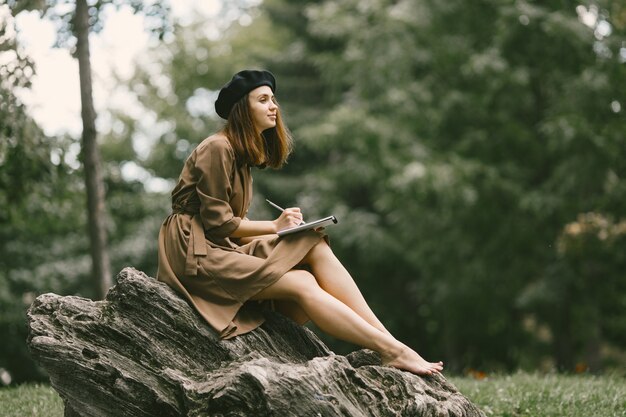 Pittrice che dipinge mentre è seduta su una roccia in un parco. Una donna che indossa un abito color cachi e un cappello nero