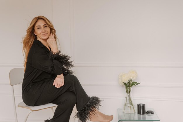 Piuttosto adulta donna caucasica bionda in pigiama nero si siede in una stanza luminosa con posto per testo Concetto di divertimento del fine settimana