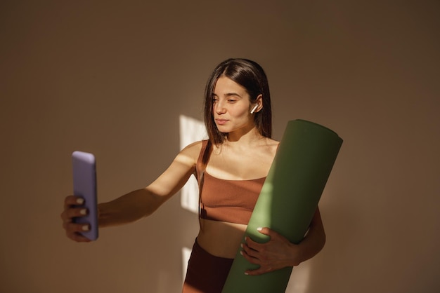 Piuttosto giovane donna caucasica con tappetino fitness prende selfie su iPhone al chiuso La donna bruna in abbigliamento sportivo gode della tecnologia moderna Concetto di stile di vita per il tempo libero