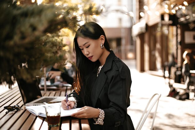 Piuttosto giovane donna in trench nero annotare le informazioni nel taccuino Donna bruna asiatica lavora nel computer portatile all'esterno