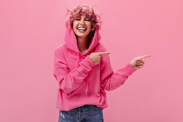 Piuttosto giovane donna riccia con i capelli rosa sorride ampiamente e guarda nella fotocamera Cute ragazza attiva indica di posizionare il testo su sfondo isolato
