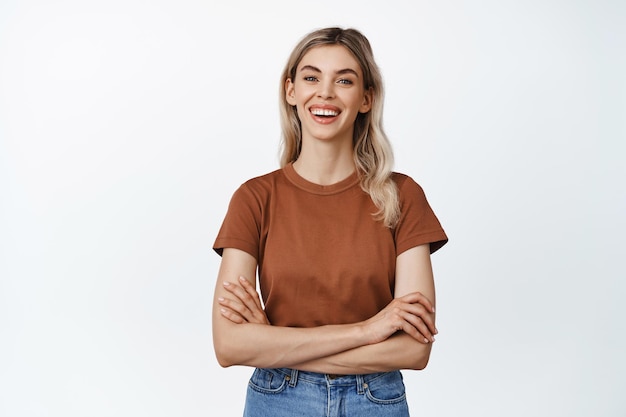 Piuttosto giovane donna ridere e sorridere incrociare le braccia sul petto e guardando la fotocamera in piedi in abiti casual su sfondo bianco