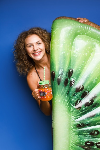 Piuttosto giovane donna vestita in costume da bagno in posa con kiwi galleggiante e cocktail su blue