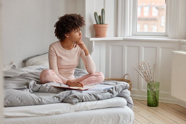 Piuttosto giovane ragazza dalla pelle scura con taglio di capelli afro, scrive sul diario, ha un'espressione sognante, indossa un pigiama, posa sul letto in una spaziosa camera da letto, è immersa nei pensieri prima di dormire. Concetto di biancheria da letto