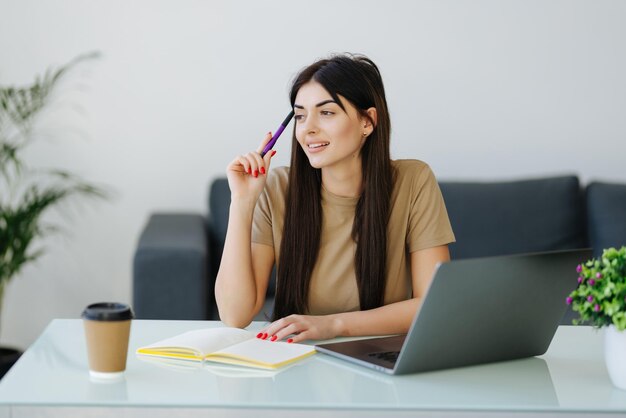 Piuttosto giovane ragazza sedersi desktop pc tenere penna scrivere blocco note indossare occhiali camicia in ufficio a casa al chiuso