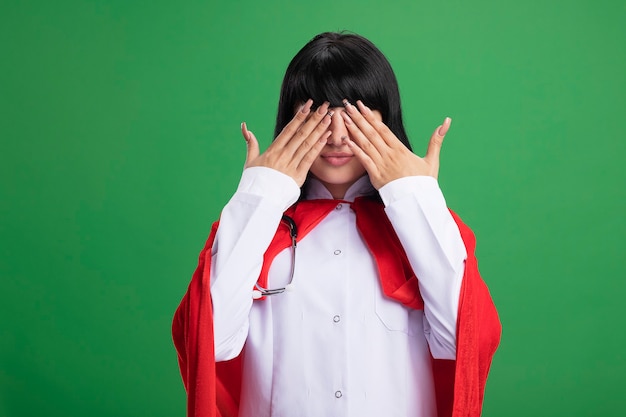 Plesased giovane ragazza supereroe che indossa uno stetoscopio con veste medica e mantello coperto gli occhi con le mani isolate sulla parete verde