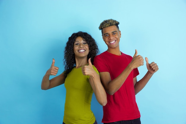 Pollice in alto, sorridente. Giovane uomo afro-americano emotivo e donna in abiti colorati su sfondo blu. Bella coppia. Concetto di emozioni umane, esperienza facciale, relazioni, annuncio, amicizia.