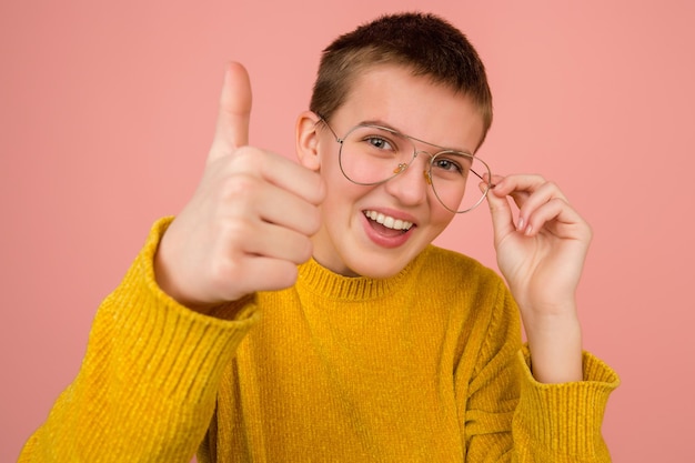 Pollice in su, sorridente. Ritratto di ragazza caucasica su sfondo rosa corallo studio con copyspace per annuncio. Bellissimo modello con gli occhiali. Concetto di emozioni umane, espressione facciale, vendite, pubblicità, moda.