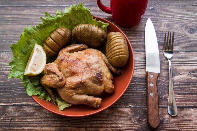 Pollo arrosto intero in una ciotola con patate; coltello e forchetta sul tavolo di legno