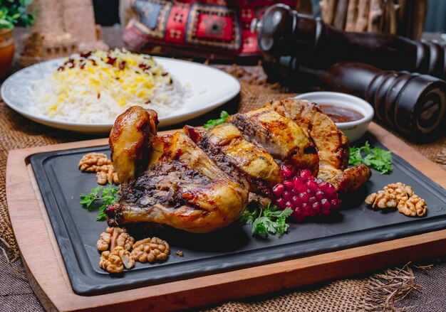 Pollo ripieno arrosto decorato con melograno e noci su un bordo nero e riso in un piatto bianco su un tavolo di legno