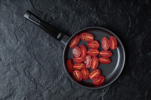 Pomodori ciliegia freschi tagliati a metà in padella nera su sfondo nero.