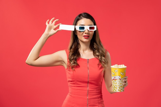 Popcorn della holding della giovane femmina di vista frontale con il biglietto in occhiali da sole di d sullo scrittorio rosso