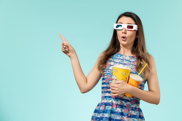 Popcorn e bevanda della tenuta della giovane femmina di vista frontale in occhiali da sole di d sullo scrittorio blu